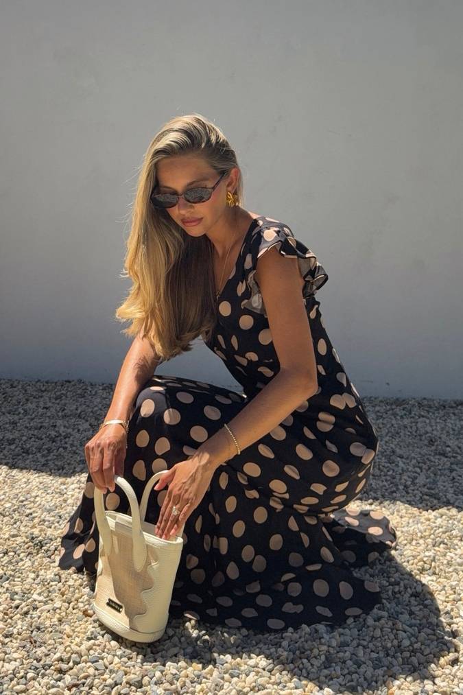 Woman in a black polka dot dress holding a beige handbag against a gray wall.