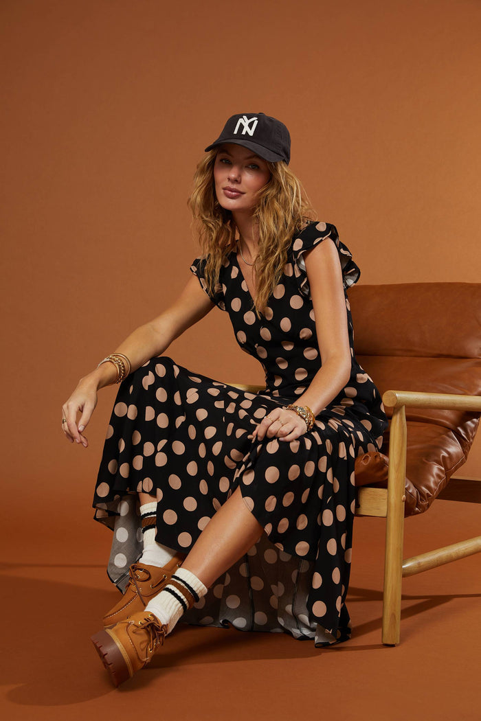 Woman in a polka dot dress and cap sitting on a brown chair against a brown background