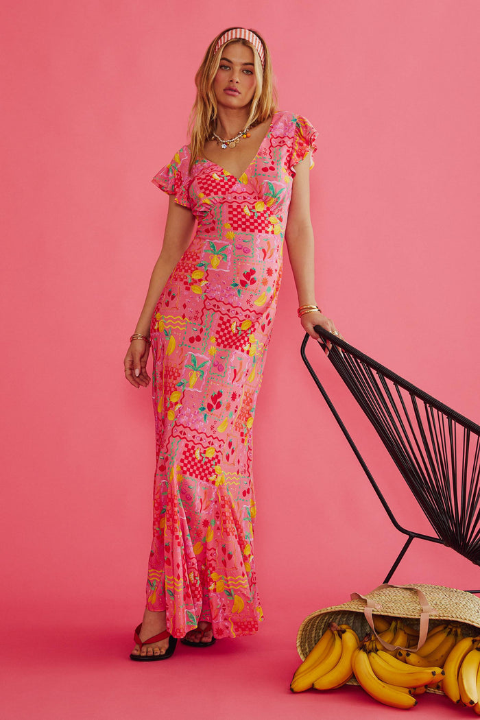 Woman in a colorful dress standing next to a black chair and bananas on a pink background