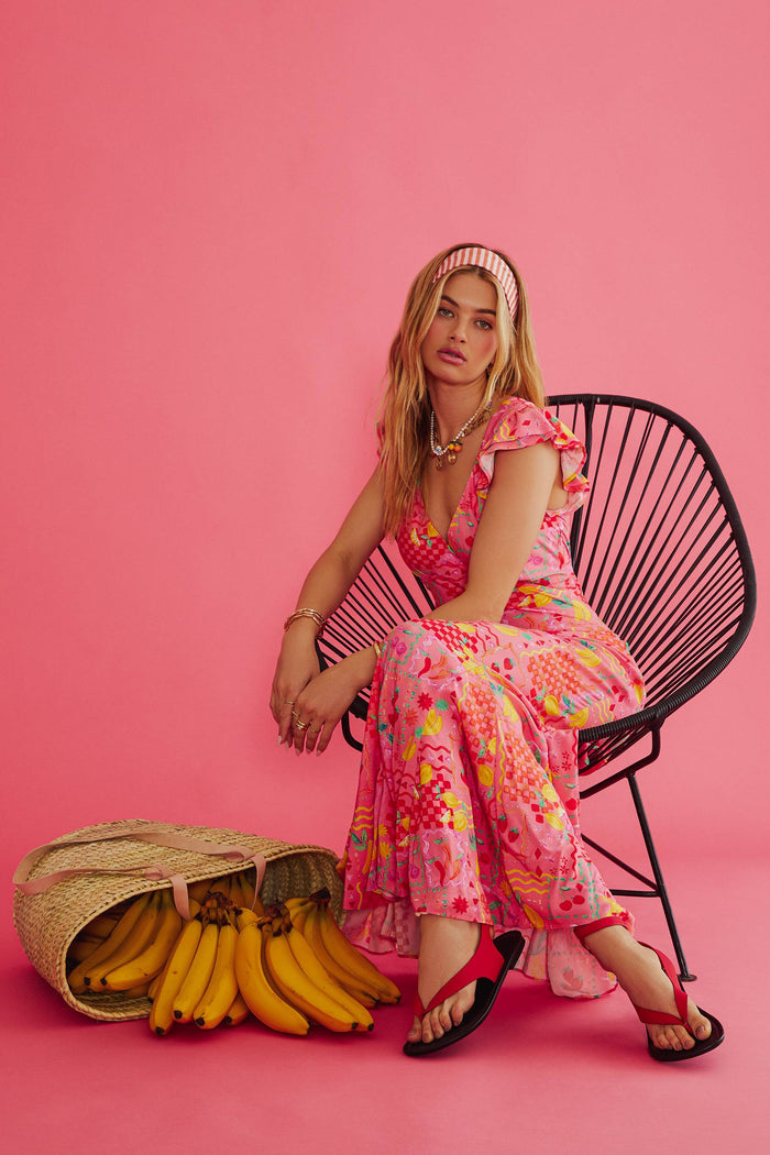 Woman in a colorful dress sitting on a chair with a basket of bananas against a pink background
