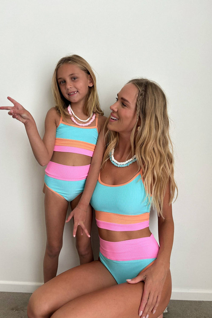 Two women in matching colorful striped swimsuits posing against a white wall.