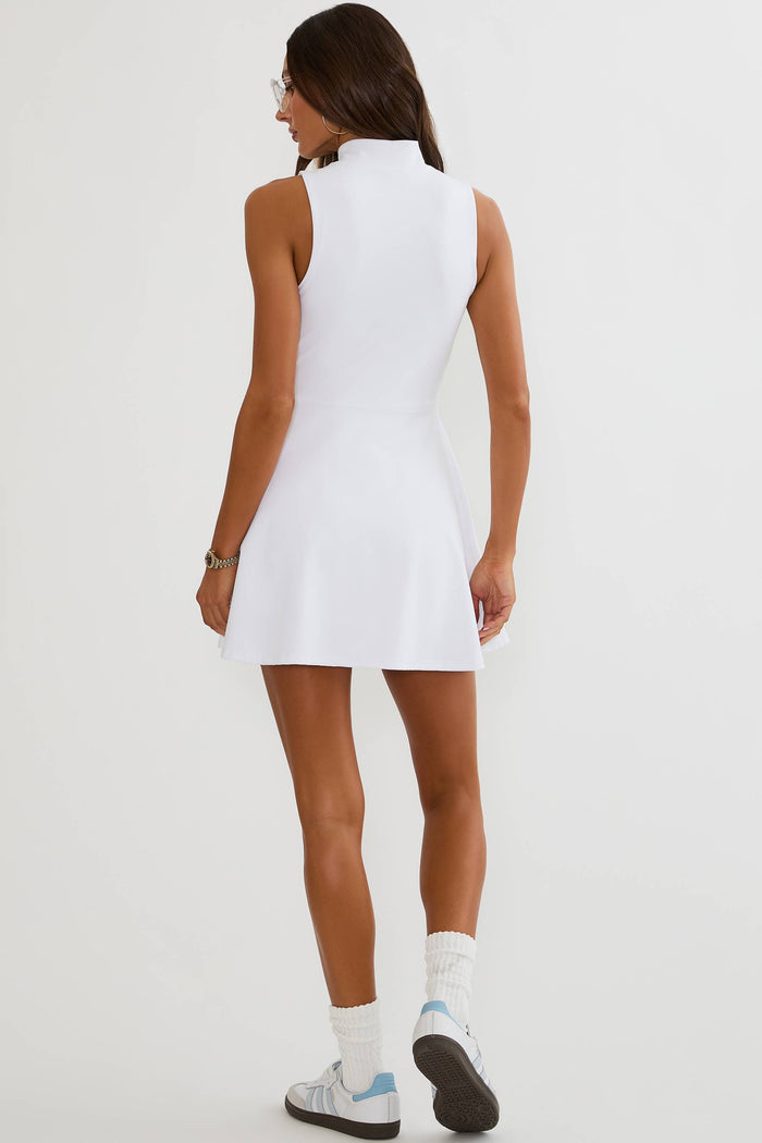 A woman shown from the back wearing a sleeveless white tennis dress, white socks, and white sneakers with light blue accents, set against a plain white studio background.