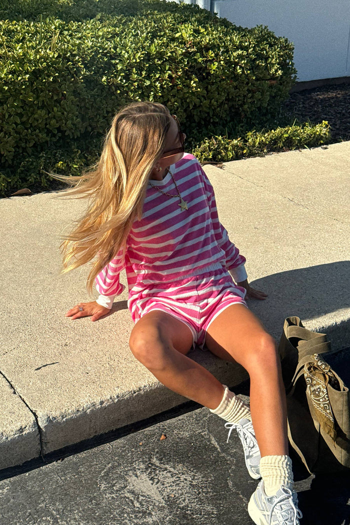Child in a pink and white striped outfit sitting on a sidewalk with green bushes in the background.