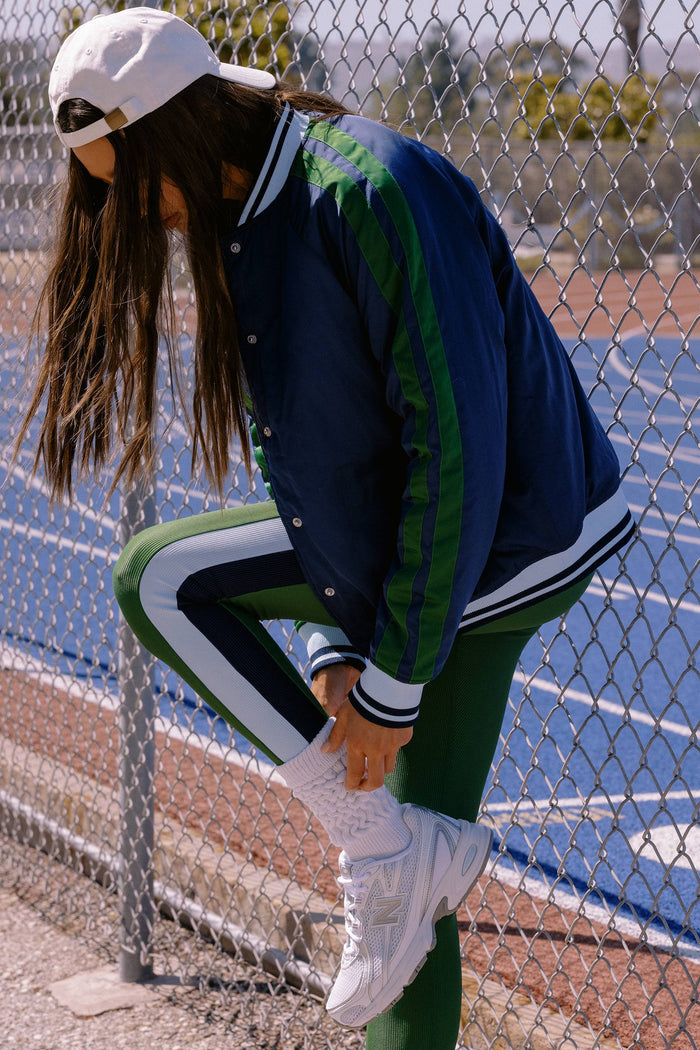 Person wearing a navy and green varsity jacket with white pants, standing on a tennis court.