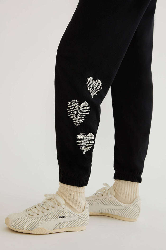 Black sweatpants with white hearts on the side worn by a woman on a plain white background.