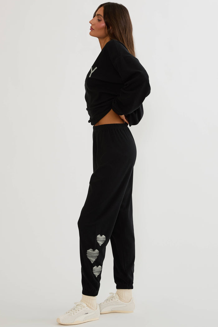 Black sweatpants with white hearts on the side worn by a woman on a plain white background.