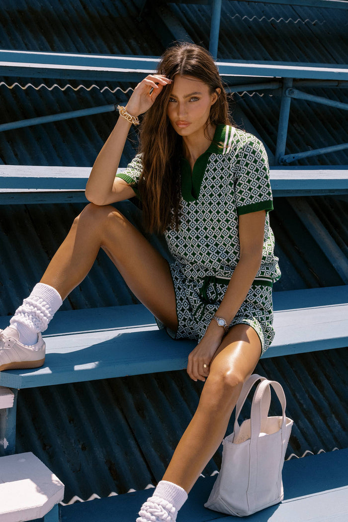 Woman sitting on a blue bench wearing a patterned outfit with white socks and a gray bag.