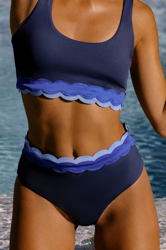 A woman wearing a two-piece swimsuit with a navy blue base and layered light blue scalloped trim, photographed outdoors near water.