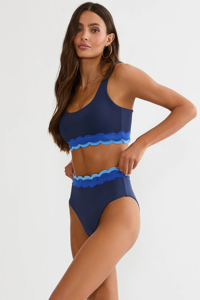 Woman wearing navy high waisted bottom with shades of blue scalloping on a white wall.