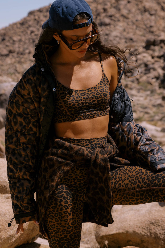 A woman sitting outdoors on rocks wearing a matching leopard-print lounge set, including a sports bra, crop top, shorts, and sweatpants, with a matching leopard-print jacket draped over her shoulders. She is also wearing a blue baseball cap and sunglasses.