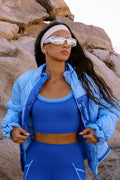 A woman wearing a blue athletic set consisting of a sports bra, leggings, and a matching headband, styled with a light blue puffer jacket and mirrored sunglasses, posing outdoors against a rocky desert background.