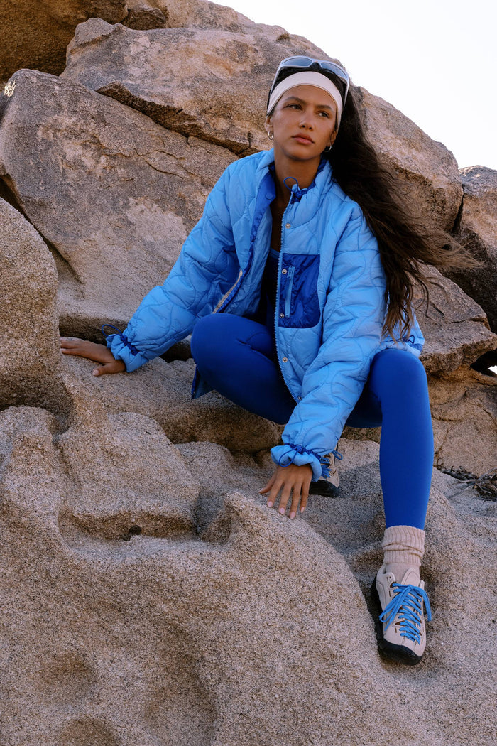 A woman sits on large tan rocks outdoors, wearing a light blue puffer jacket, bright blue leggings, white socks, blue and white sneakers, and a white headband with sunglasses.