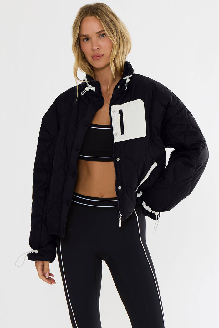 A woman modeling a black puffer jacket with a large white chest pocket featuring a black number '1' and white piping details, paired with black athletic leggings and a black striped crop top against a light gray studio background.