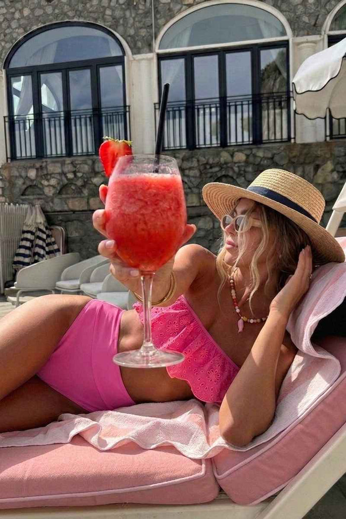 Woman in pink bikini and sun hat holding a red cocktail on a sun lounger.