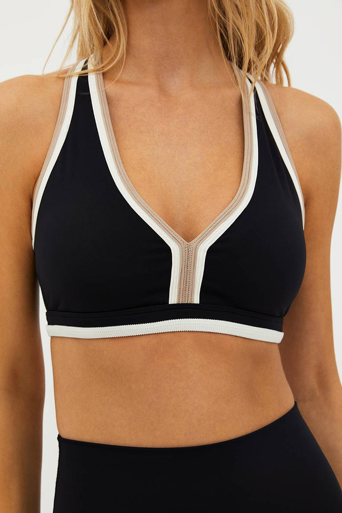A woman wearing a black sports bra with color-blocked binding in tan and white, featuring a plunge neckline and racerback detail.