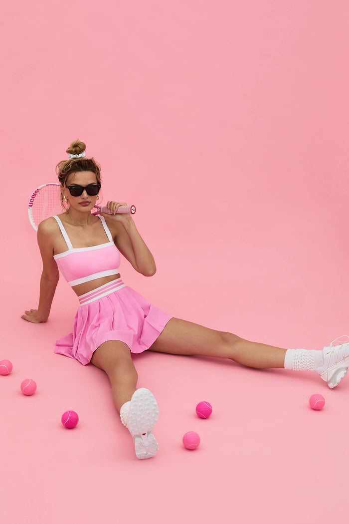 pink pleated tennis skirt with pink and white contrast trim and a pink sports bra white white straps and a white contrast trim