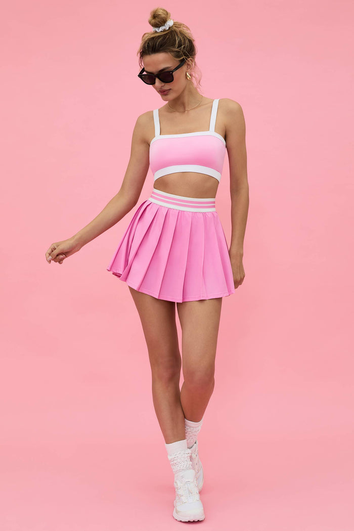 pink pleated tennis skirt with pink and white contrast trim and a pink sports bra white white straps and a white contrast trim