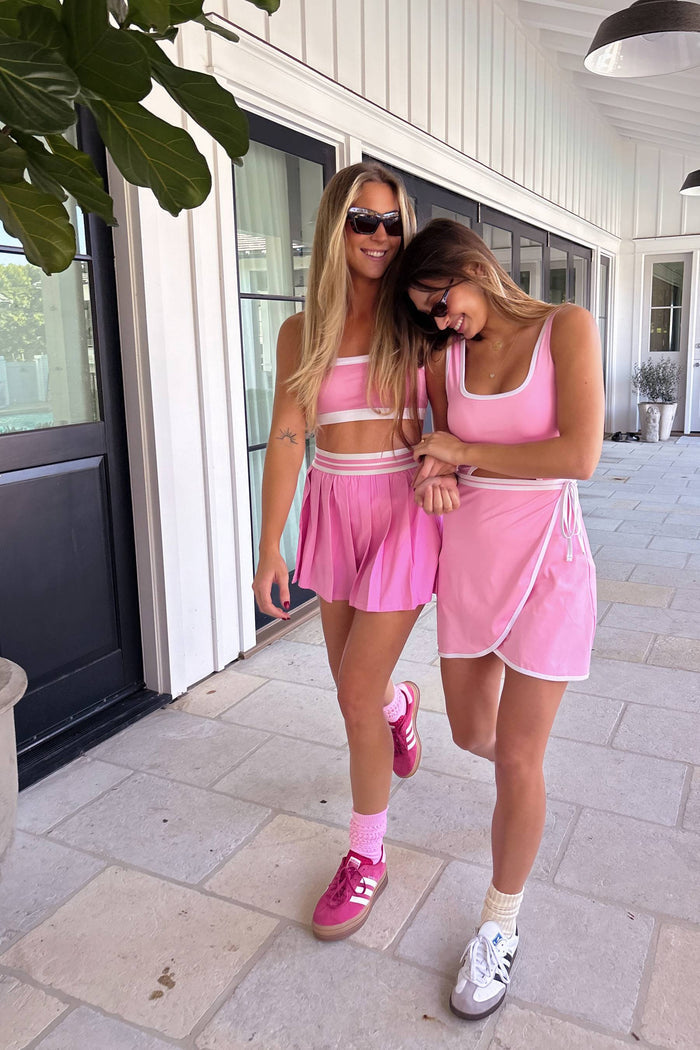 pink pleated tennis skirt with pink and white contrast trim and a pink sports bra white white straps and a white contrast trim