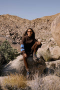 A woman wearing a dark brown knit sweater with light blue and white sleeve stripes, brown leggings, and glasses sits on a large rock in a rocky desert landscape.