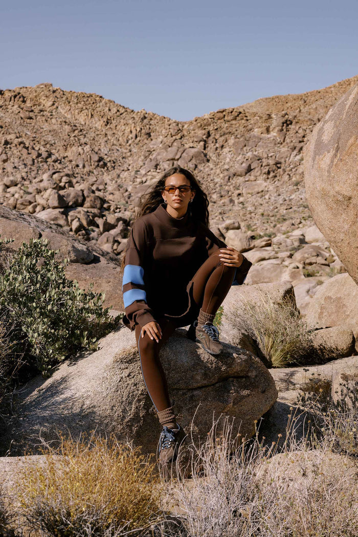 A woman wearing a dark brown knit sweater with light blue and white sleeve stripes, brown leggings, and glasses sits on a large rock in a rocky desert landscape.
