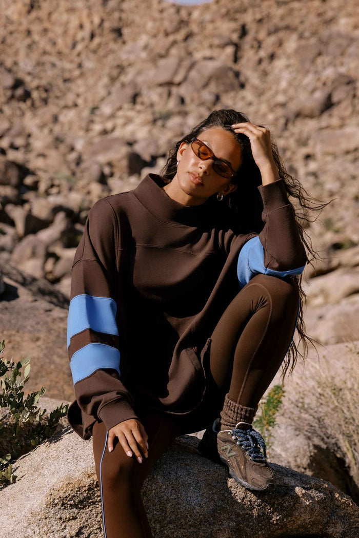 A woman wearing a brown mock-neck sweatshirt with light blue color-blocked sleeves and matching brown leggings, sitting on a rock in a rocky outdoor environment.