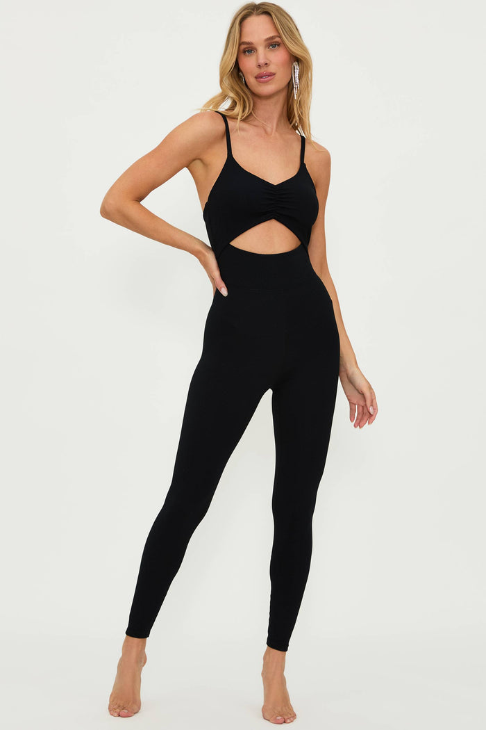 A female model wearing a black catsuit with a deep V-neckline, cutout detail, and adjustable straps.