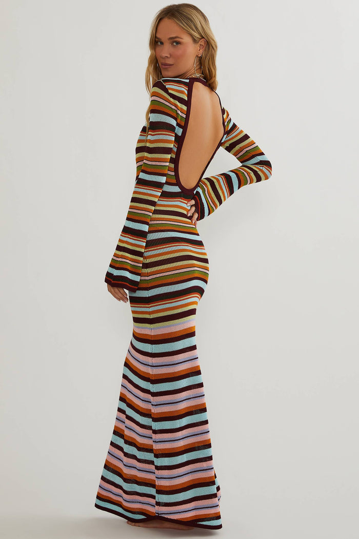 A woman modeling a long-sleeve maxi dress with a multicolor horizontal stripe pattern, featuring a dramatic open back and flared hem, set against a plain light gray studio background.