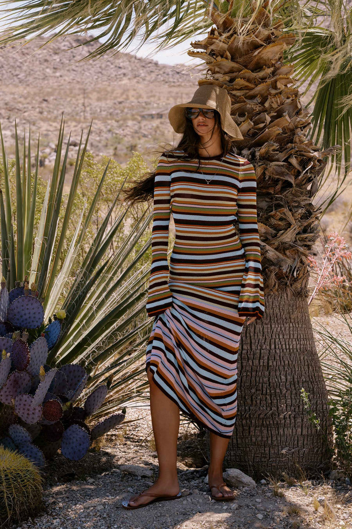 A woman wearing a long-sleeve striped midi dress, a wide-brimmed straw hat, and sunglasses stands outdoors next to a palm tree in a desert landscape with cacti.