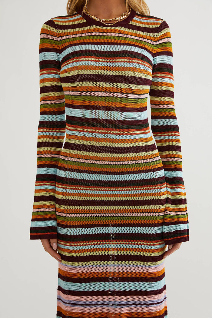 A woman wearing a long-sleeve ribbed knit midi dress with horizontal multicolor stripes in brown, orange, yellow, green, light blue, and cream, shown against a plain light gray studio background.
