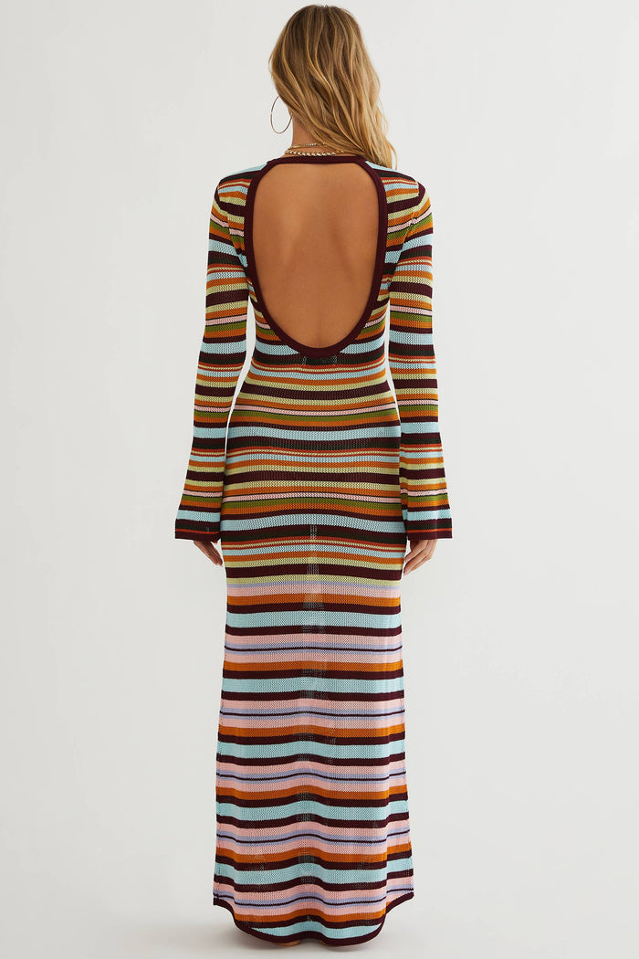 A woman viewed from the back wearing a long-sleeve maxi dress with a large open back and horizontal multicolor stripes in orange, yellow, brown, blue, and white, against a plain light gray studio background.