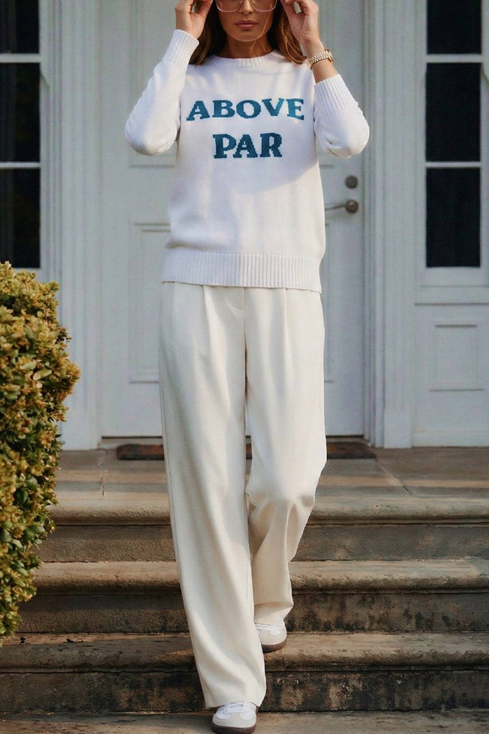 Person wearing a white sweater with 'ABOVE PAR' text and white pants standing on steps.