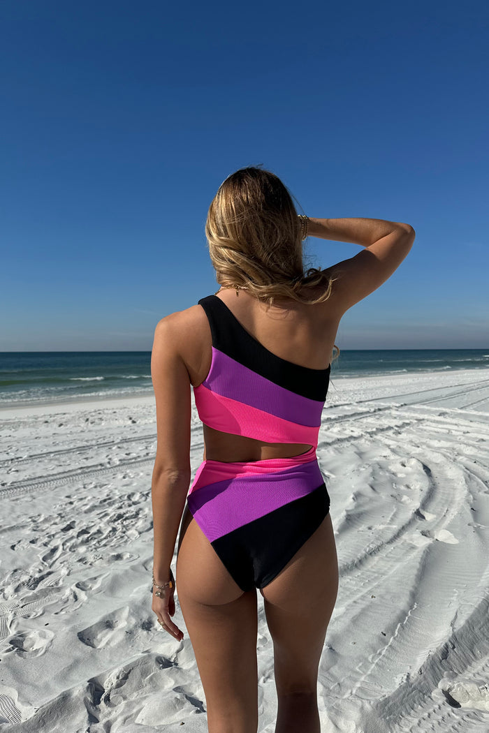 A model wearing a one-piece swimsuit with color blocking in vivid violet, featuring neon pink, glowing purple, and sleek black colors, with a side cutout.