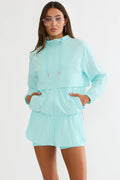 A woman modeling a matching mint green athletic set consisting of a lightweight hooded jacket and coordinating shorts, with pink drawstring tips and pink zipper accents.