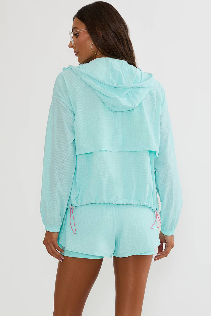 A woman shown from the back wearing a light mint green hooded windbreaker jacket and matching shorts against a plain light gray studio background.