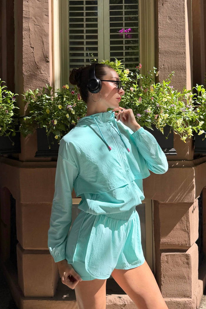 A woman wearing a matching mint green athletic tracksuit, consisting of a lightweight zip-up jacket and matching shorts, posing outdoors in front of a stone building with potted plants.