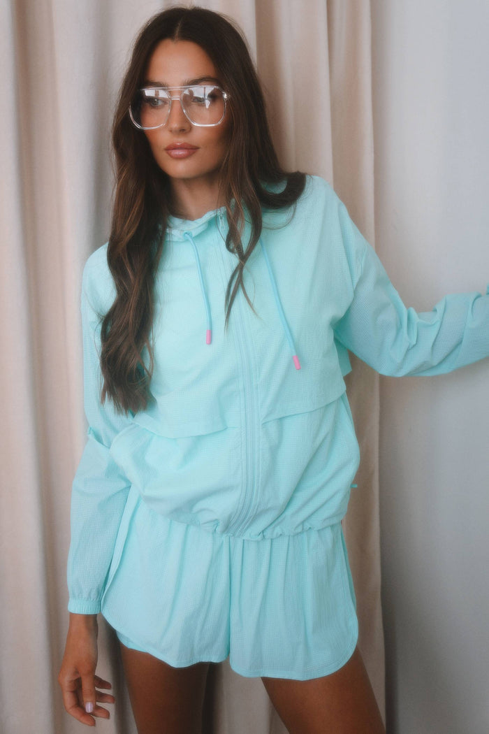 A woman wearing a light mint green hooded long-sleeve romper with a front zipper and drawstrings, posed against a neutral beige curtain background.