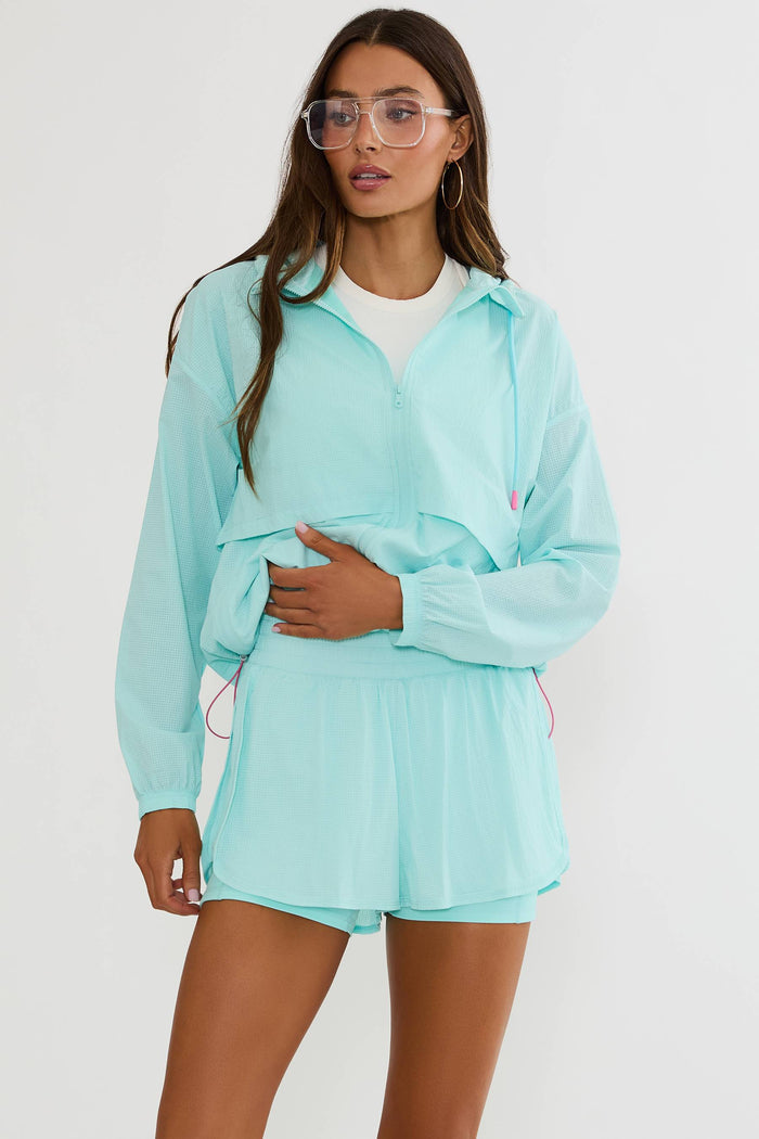 A woman modeling a light aqua two-piece athletic set consisting of a long-sleeve zip-up windbreaker jacket and matching shorts, worn over a white t-shirt. She is wearing clear-framed glasses and standing against a plain white studio background.