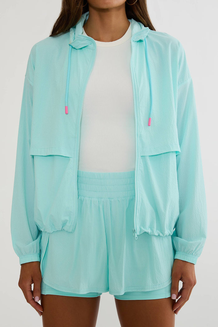 A model wearing a light mint green lightweight zip-up hooded jacket over a white top and matching mint green shorts, against a plain light gray studio background.