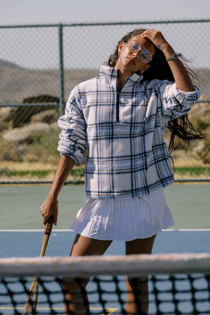 A woman on an outdoor tennis court wearing a white and blue plaid quarter-zip fleece pullover and a white tennis skirt, holding a tennis racket.
