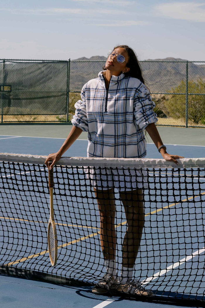 A woman stands on an outdoor tennis court behind the net, wearing a white and navy plaid quarter-zip pullover, white shorts, and holding a tennis racket.