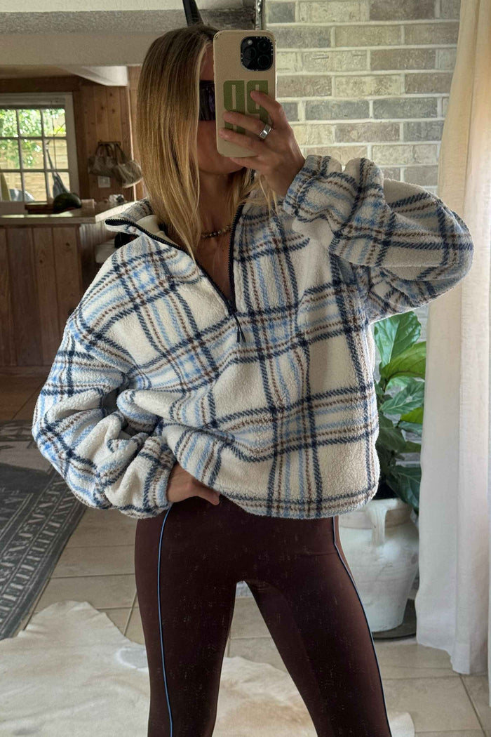 A woman takes a mirror selfie wearing a cream-colored fleece pullover with a blue and gray plaid pattern, paired with dark brown leggings.
