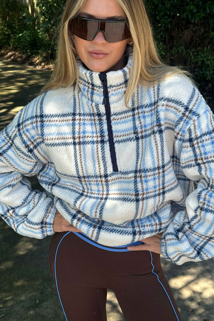 A woman outdoors wearing a light blue, white, and black plaid fleece quarter-zip pullover sweater, paired with dark brown leggings with a light blue stripe.