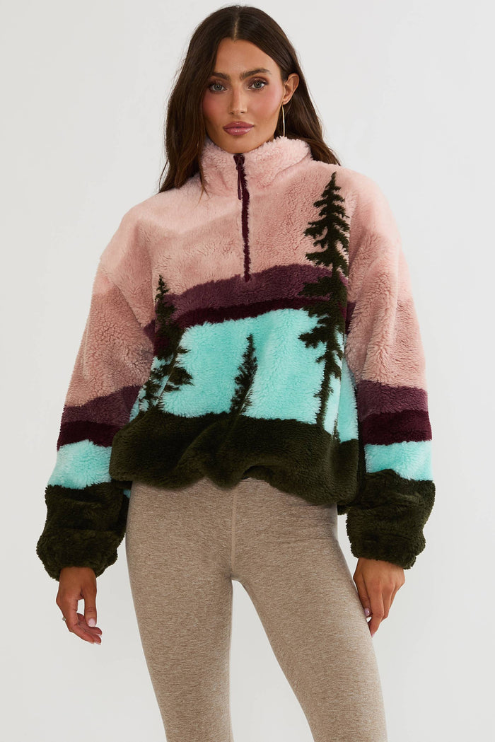 A woman modeling a fuzzy quarter-zip fleece pullover with a scenic landscape print in pink, mint green, dark green, and purple, paired with heather gray leggings against a plain white studio background.