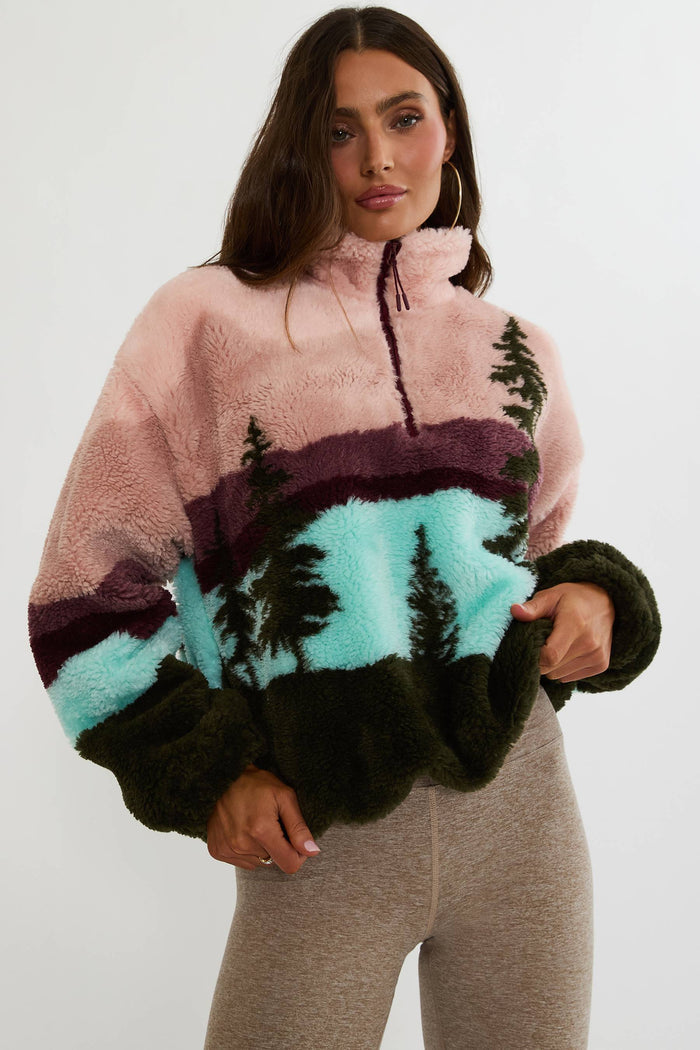 A woman wearing a fuzzy quarter-zip fleece pullover with a scenic mountain landscape print in pink, purple, turquoise, and dark green, paired with heather gray leggings, against a plain white studio background.