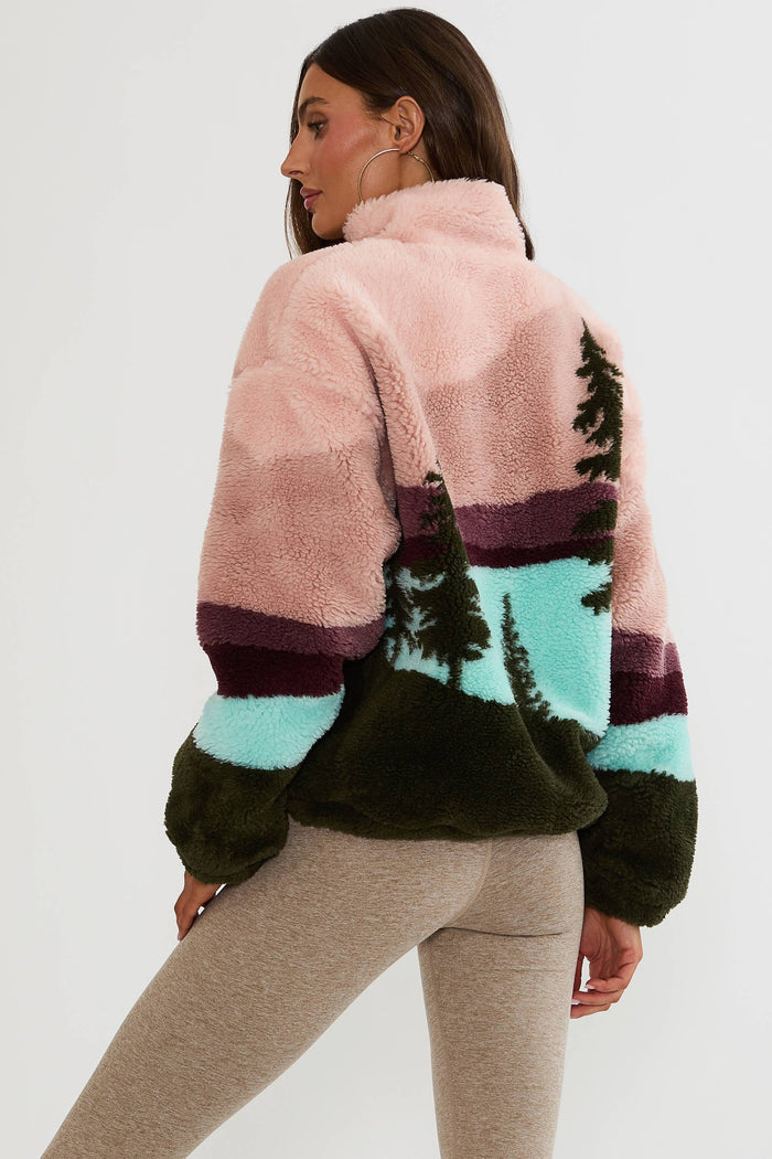 A woman shown from the back wearing a fuzzy sherpa fleece pullover with a multicolor landscape print in shades of pink, teal, dark green, and burgundy, paired with heather gray leggings.