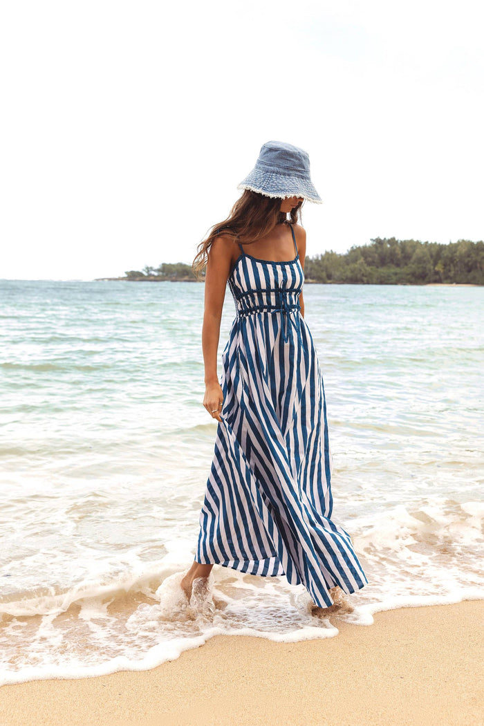 Woman in a blue and white striped dress and hat standing on a beach.