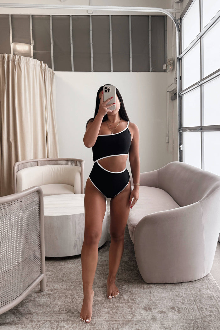 Woman taking a mirror selfie wearing a black bikini in a modern room with neutral decor.