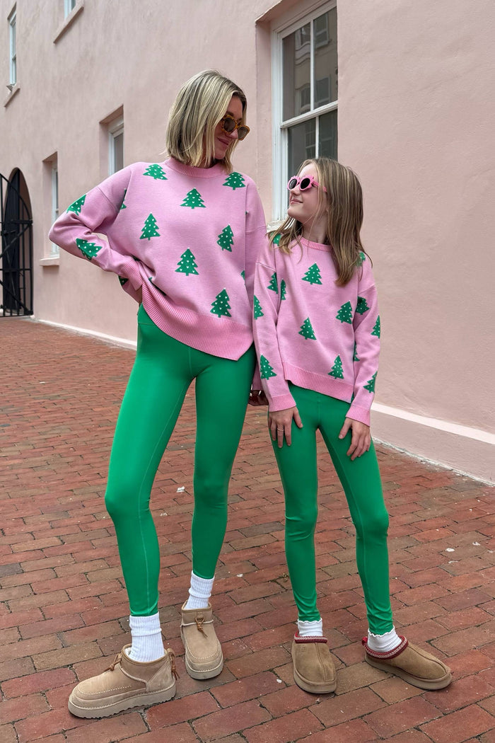 Two people wearing matching pink sweaters with green tree patterns and green leggings, standing on a brick sidewalk.