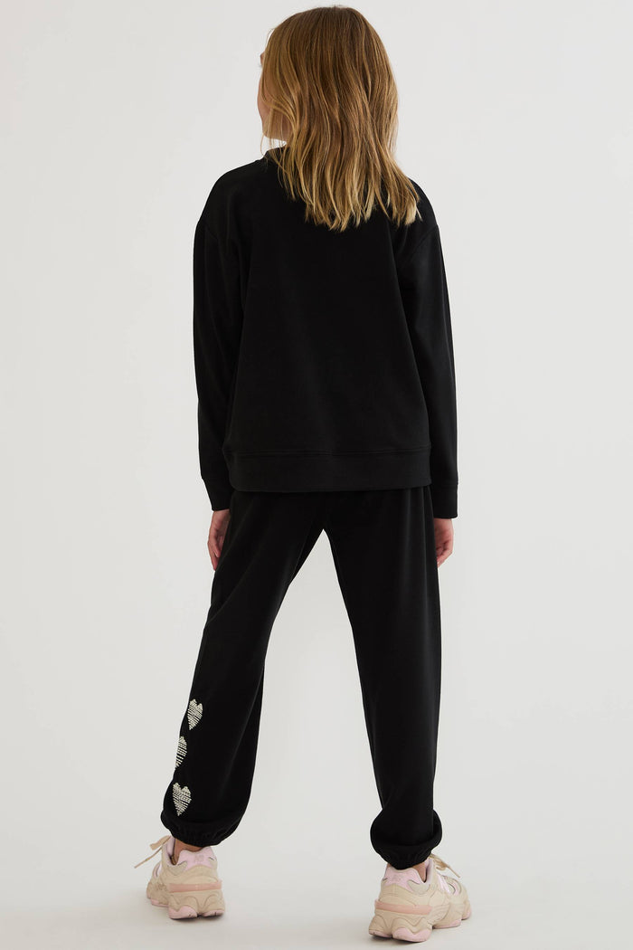 A child model shown from the back wearing a black sweatshirt and black jogger pants with small white heart details near the cuffs, paired with light pink sneakers, against a plain light gray studio background.