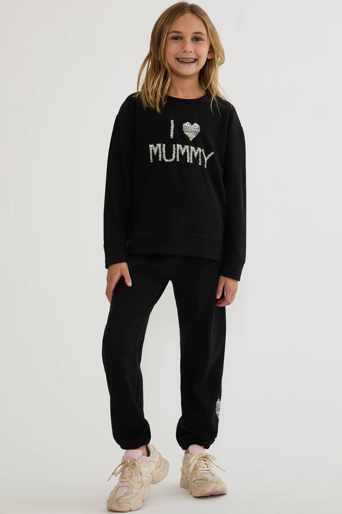 A young girl modeling a black sweatshirt and matching sweatpants set. The sweatshirt features 'I [heart] MUMMY' printed in white across the chest.